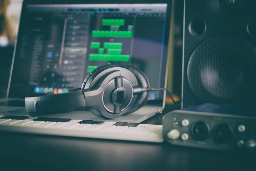 A pair of headphones sits on a laptop with music editing software on the screen. Also pictured is a music keyboard and speaker.