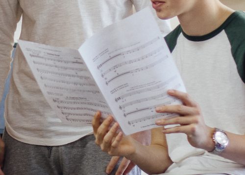 Male teenagers are in a music lesson at school. They are discussing sheet music with their teacher.