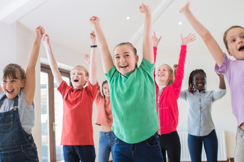 A group of children stand with their arms in the air. They are brightly dressed and smiling.