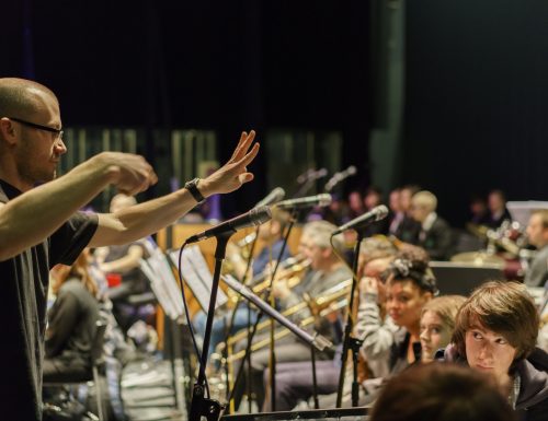 James Raywood, a pale skinned man with stubble, shaved head and glasses, conducts an orchestra of young people