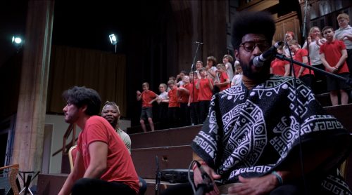 A dark skinned man, medium skinned and dark skinned woman are positioned at the bottom of some steps. They look to be singing and performing. A group of school children in red uniform stand behind them, also singing.