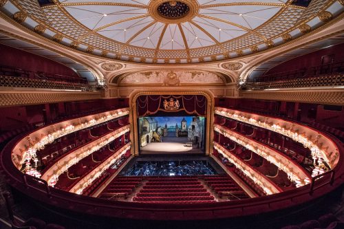 A view of the Royal Opera House from the back stalls. It is empty. The orchestra pit can be seen and the stage has a set that looks like a town at dusk.