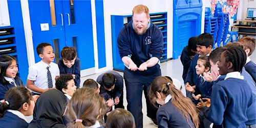 A man with light skin, red hair and a red beard stands with his palms out in the middle of a group of school children. They all look excited and ae wearing blue uniform.