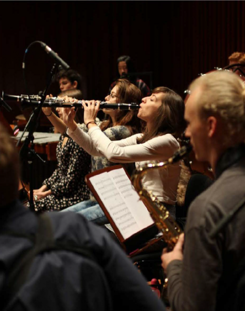 A young woman who is light skinned with dark hair, plays clarinet. She is surrounded by other musicians who are softly blurred out of focus.