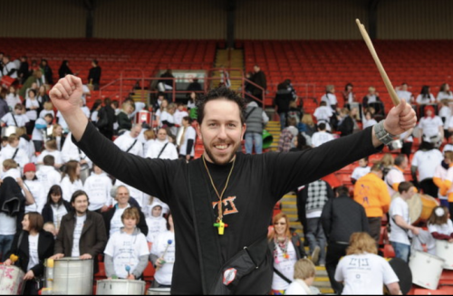 Tim brain, a pale skinned man with dark hair and a beard, stands in front of a crowd with a wide smile. He is wearing a black top, holds a drumstick and both arms are up in the air.