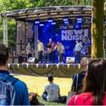 A live band performs on an outdoor stage with a "NEW NOISE" banner, surrounded by trees. Audience members, some seated on the grass and others standing, watch the performance in the sunshine.