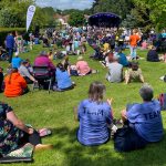 A lively outdoor festival with a crowd sitting on the grass, watching a performance on a stage. Two people in blue "TEAM" shirts sit in the foreground. The weather is sunny, and attendees are relaxed, enjoying the event.