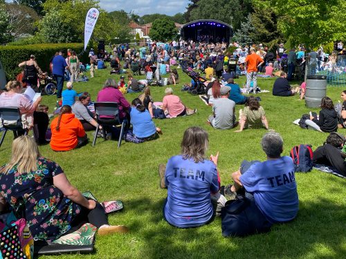 A lively outdoor festival with a crowd sitting on the grass, watching a performance on a stage. Two people in blue "TEAM" shirts sit in the foreground. The weather is sunny, and attendees are relaxed, enjoying the event.