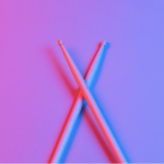 A pair of wooden drumsticks crossed over each other on a vibrant background with a gradient of pink and blue lighting.