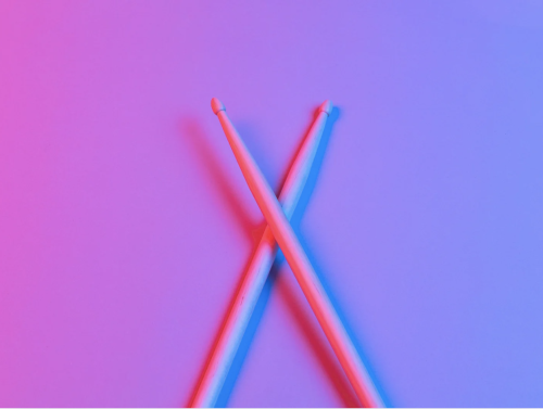 A pair of wooden drumsticks crossed over each other on a vibrant background with a gradient of pink and blue lighting.